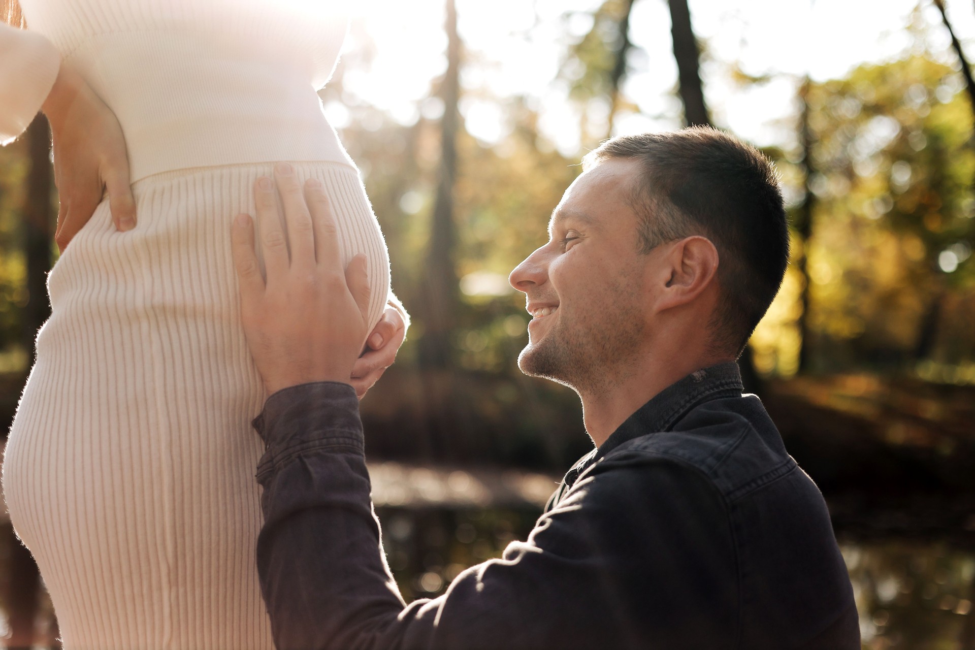 Cropped shot of happy young husband enjoying strokes, talks and kisses tummy of wife, waiting for birth baby outdoors in sunbeams in the city park. Maternity prenatal care and woman pregnancy concept.